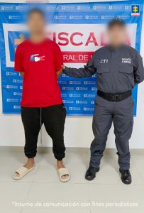 En la fotografía aparece un hombre alto, joven, de pie con los brazos atrás, trigueño, de contextura gruesa, con cabellos negros, vestido con camiseta roja, sudadera negra y chanclas beige. Está custodiado por un uniformado del CTI. Detrás de ellos hay una bandera azul de la Fiscalía General.