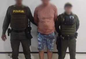 En la fotografía está un hombre alto, trigueño, se encuentra de pie, con los brazos atrás, rostro difuso, vestido con una camiseta rosada de bermuda de jean y sandalias rosadas., custodiado por dos uniformados de la Policía Nacional.