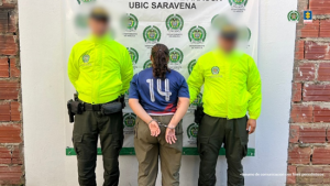 Se visualiza a la capturada de espaldas junto a dos uniformados de la Policía Nacional. Detrás el banner institucional de la Policía Nacional