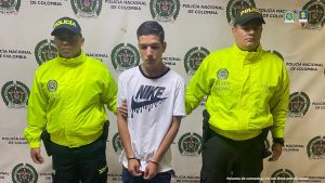 El detenido aparece de frente con una camiseta blanca y esposado, acompañado de dos uniformados de la Policía Nacional