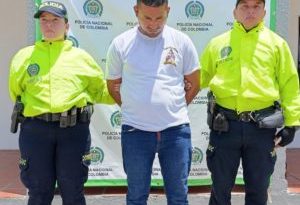 En la fotografía aparece una persona capturada, acompañado de un uniformado de la Policía Nacional y del Ejército Nacional.  En la parte posterior un banner con logos de la entidad.