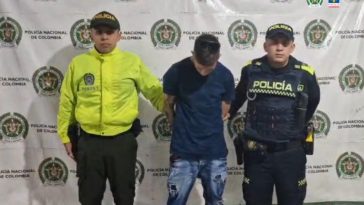 El procesado se encuentra en custodia por dos uniformados de la Policía Nacional. El capturado tiene un buzo color azul oscuro y un jean. Detrás se observa un backing de la Policía.