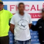 En la fotografía aparece un hombre de pie, con el rostro difuso, alto moreno, vestido con camiseta blanca, custodiado por uniformados del CTI y de la Policía Nacional. Detrás de ellos hay un pendón azul de la Fiscalía.