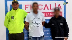 En la fotografía aparece un hombre de pie, con el rostro difuso, alto moreno, vestido con camiseta blanca, custodiado por uniformados del CTI y de la Policía Nacional. Detrás de ellos hay un pendón azul de la Fiscalía.