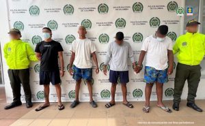 En la fotografía aparecen cuatro hombres de pie, con los brazos esposados, todos vestidos con camisetas y bermudas, custodiados por dos uniformados de la Policía Nacional.