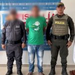 En la fotografía está un hombre de pie con el rostro difuso, con los brazos atrás, vestido con una camiseta verde, un jean azul y sandalias negras. Se encuentra acompañado por un uniformado del CTI y un uniformado de la Policía Nacional.