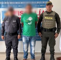 En la fotografía está un hombre de pie con el rostro difuso, con los brazos atrás, vestido con una camiseta verde, un jean azul y sandalias negras. Se encuentra acompañado por un uniformado del CTI y un uniformado de la Policía Nacional.