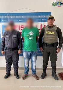 En la fotografía está un hombre de pie con el rostro difuso, con los brazos atrás, vestido con una camiseta verde, un jean azul y sandalias negras. Se encuentra acompañado por un uniformado del CTI y un uniformado de la Policía Nacional.