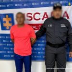 En la fotografía aparece un hombre moreno de pie con los brazos atrás, vestido con camiseta roja y pantalón azul, custodiado por un uniformado del CTI. Detrás de ellos hay un pendón de la Fiscalía General.