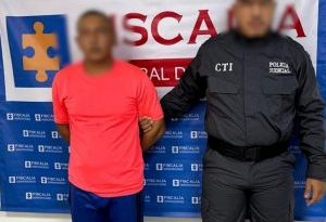 En la fotografía aparece un hombre moreno de pie con los brazos atrás, vestido con camiseta roja y pantalón azul, custodiado por un uniformado del CTI. Detrás de ellos hay un pendón de la Fiscalía General.