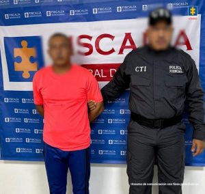 En la fotografía aparece un hombre moreno de pie con los brazos atrás, vestido con camiseta roja y pantalón azul, custodiado por un uniformado del CTI. Detrás de ellos hay un pendón de la Fiscalía General.