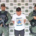 El imputado se encuentra custodiado por dos uniformados de la Policía Nacional. El capturado tiene un buzo blanco. Detrás se observa un backing de la Policía.