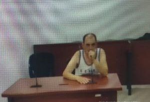 En la fotografía aparece un hombre blanco, semi calvo, con la mano puesta en la boca, vestido con una camisilla blanca y se encuentra sentado en un escritorio de madera con un micrófono, en la sala de audiencia donde se le procesó.