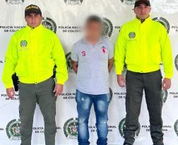 El procesado se encuentra custodiado por dos uniformados de la Policía Nacional. Detrás se observa un backing de la Policía. El agresor tiene cubierto el rostro.