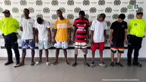 En la fotografía están cinco hombres de pie, esposados, de frente, con las cabezas agachadas, todos vestidos con camisetas y bermudas. Se encuentran custodiado por dos uniformados de la Policía Nacional.