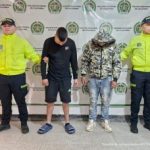 Los imputados se encuentran bajo custodia de dos uniformados de la Policía Nacional.  Uno de los capturados vestía pantalón corto negro y una chaqueta, mientras que el otro vestía jeans con una chaqueta con capucha.  Detrás se puede ver el apoyo policial.