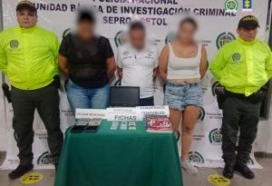 en la imagen se ven tres personas detenidas bajo custodia de dos integrantes de la Policía Nacional. Detrás suyo un backing institucional, delante de ellos una mesa con elementos incautados.