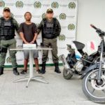 En la fotografía un hombre moreno, de pie, de estatura baja, con los brazos adelantes esposados, vestido con una camiseta negra, bermuda gris, tenis negros, custodiado por dos uniformados de la Policía Nacional.