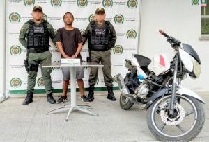 En la fotografía un hombre moreno, de pie, de estatura baja, con los brazos adelantes esposados, vestido con una camiseta negra, bermuda gris, tenis negros, custodiado por dos uniformados de la Policía Nacional.