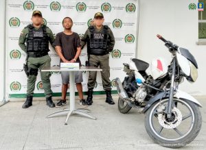 En la fotografía un hombre moreno, de pie, de estatura baja, con los brazos adelantes esposados, vestido con una camiseta negra, bermuda gris, tenis negros, custodiado por dos uniformados de la Policía Nacional.