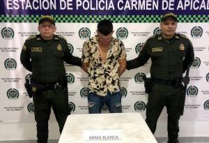 en la imagen se ve una persona detenida bajo custodia de dos uniformados de la Policía. Detrás suyo un backing institucional.