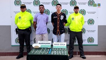 en la fotografia se visualiza a los dos detenidos en medio de dos funcionarios de la policia, en frente de ellos se ve una mesa donde se encuentran los estupefacientes y detras de ellos se ve un baking de la policia