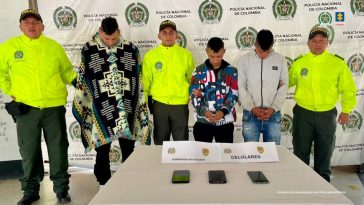 Los capturados se encuentran de frente y custodiados por tres uniformados de la Policía Nacional. Detrás se observa un backing de la Policía Nacional. En la foto se ve una mesa con tres celulares incautados.