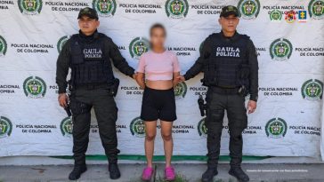 en la imagen se ve una mujer detenida bajo custodia de dos integrantes de la Policía. Detrás suyo un backing institucional.