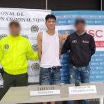 Se visualiza al capturado junto a un uniformado de la Policía Nacional y un investigador del CTI. Frente a ellos una mesa con los elementos incautados: una granada de fragmentación y munición. Detrás de ellos los banners que identifican a la Policía Nacional y a la Fiscalía General de la Nación