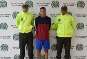 En la fotografía aparece un hombre de pie, delgado, con los brazos atrás, moreno, de cabellos cortos crespos negros, vestido con un buzo rojo, bermuda azul. Se encuentra custodiado por dos uniformados de la Policía Nacional.