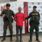 Se observa al capturado custodiado por dos uniformados de la Policía Nacional. En la imágen se puede apreciar de fondo un backing de la Policía y el Ejército Nacional. El capturado tiene una camisa color roja.