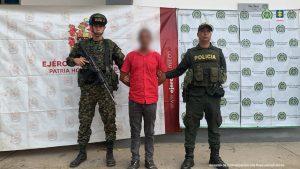 Se observa al capturado custodiado por dos uniformados de la Policía Nacional. En la imágen se puede apreciar de fondo un backing de la Policía y el Ejército Nacional. El capturado tiene una camisa color roja.