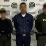 Se observa al judicializado en custodia por dos uniformados de la Policía Nacional. El rostro del capturado y de los policías se encuentran cubiertos. Detrás se encuentra un backing de la Policía.