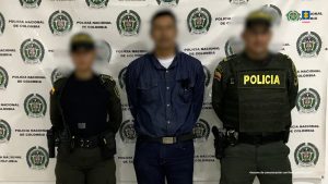 Se observa al judicializado en custodia por dos uniformados de la Policía Nacional. El rostro del capturado y de los policías se encuentran cubiertos. Detrás se encuentra un backing de la Policía.