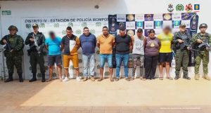 En la fotografía aparecen varios hombres de pie, parados con los brazos atrás, custodiados por uniformados de la Policía y la Armada Nacional. Detrás de ellos hay  pendones de esas instituciones y de la Fiscalía.