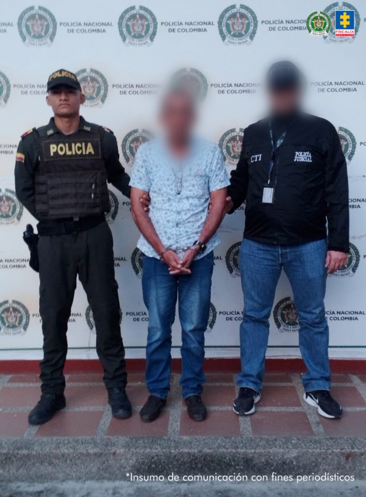 En la fotografía aparece un hombre de pie, de mediana estatura, con el rostro difuso, las manos adelantes esposadas, vestido con una camisa azul clara, un jean azul oscuro y zapatos negros. Se encuentra custodiado por un uniformado del CTI y uno de la Policía Nacional.