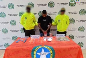 En la fotografía aparece un hombre moreno de pie, con los brazos adelante esposados, cabellos crespos negros, vestido con una camiseta negra y jean azul, custodiado por dos uniformados de la Policía Nacional.