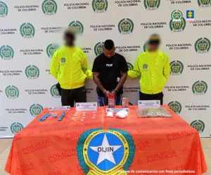 En la fotografía aparece un hombre moreno de pie, con los brazos adelante esposados, cabellos crespos negros, vestido con una camiseta negra y jean azul, custodiado por dos uniformados de la Policía Nacional.