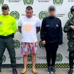 En la fotografía se aprecia de pie y con los brazos atrás al imputado. Viste camiseta blanca, de manga corta y con estampado circular en color naranja; bermuda de colores y pantuflas. Al lado derecho de él hay un uniformado de la Policía Nacional y al izquierdo un uniformado del Ejército. Detrás de ellos hay un pendón institucional de la Policía.