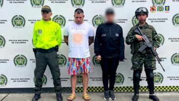 En la fotografía se aprecia de pie y con los brazos atrás al imputado. Viste camiseta blanca, de manga corta y con estampado circular en color naranja; bermuda de colores y pantuflas. Al lado derecho de él hay un uniformado de la Policía Nacional y al izquierdo un uniformado del Ejército. Detrás de ellos hay un pendón institucional de la Policía.