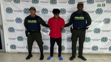 En la fotografía se aprecia de pie y con los brazos atrás  a Carlos Manuel Quiñónez. Vista camisa de manga corta de color rojo, pantalón negro y zapatillas azules. A sus extremos hay uniformados de la Policía Nacional. Detrás de ellos hay un pendón institucional de la Policía.