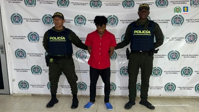 En la fotografía se aprecia de pie y con los brazos atrás  a Carlos Manuel Quiñónez. Vista camisa de manga corta de color rojo, pantalón negro y zapatillas azules. A sus extremos hay uniformados de la Policía Nacional. Detrás de ellos hay un pendón institucional de la Policía.