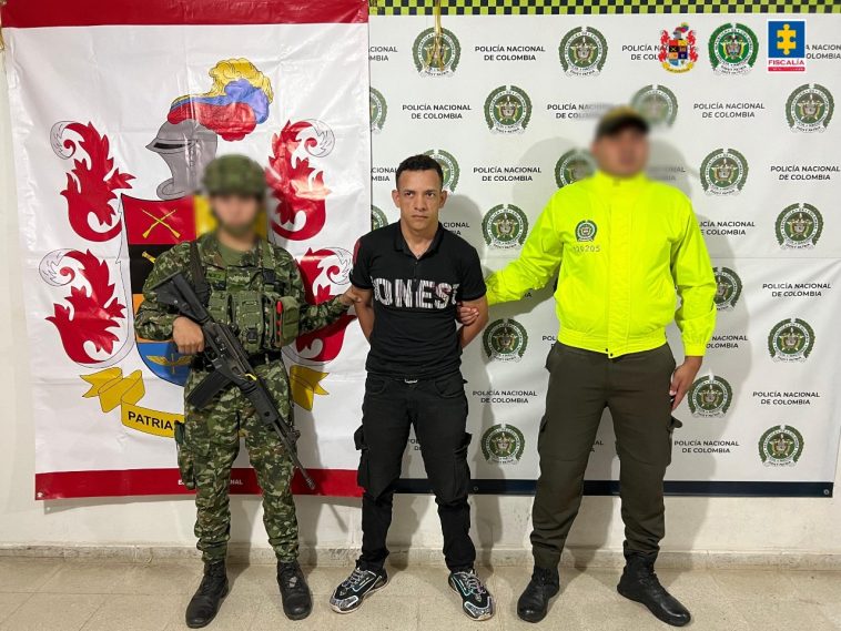 El capturado aparece en el centro de la imagen, con un miembro del Ejército en uno de sus costados y en el otro uno de la Policía. Al fondo aparecen banner de las dos instituciones.