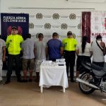 en la imagen se ve un grupo de personas detenidas bajo custodia de integrantes de la Policía, CTI de la Fiscalía y Ejército. Detrás suyo backings institucionales. Delante de ellos una moto y una mesa con elementos incautados.