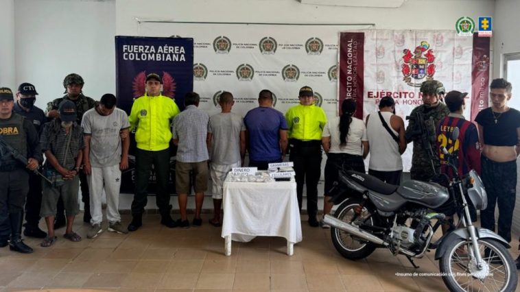 en la imagen se ve un grupo de personas detenidas bajo custodia de integrantes de la Policía, CTI de la Fiscalía y Ejército. Detrás suyo backings institucionales. Delante de ellos una moto y una mesa con elementos incautados.