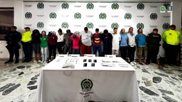 En la imagen se ve un grupo de personas detenidas bajo custodia de la Policía Nacional. Delante de ellos una mesa con elementos incautados. Detrás suyo un backing institucional.