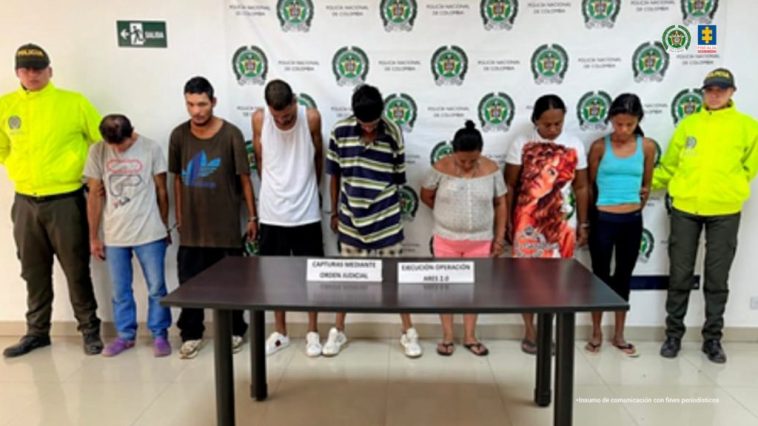 En la fotografía se aprecia a 7 de los 8 presuntos integrantes del grupo delincuencial Nueva Generación Ares 2.0. Hay cuatro hombres y tres mujeres. A sus extremos hay uniformados de la Policía Nacional. Delante de ellos hay una mesa color marrón y sobre ella hay dos letreros. Detrás del grupo hay un pendón institucional de la Policía.