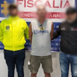 Se visualiza al capturado con el rostro difuminado, junto a un uniformado de la Policía Nacional e investigador del CTI. Detrás de ellos el banner que identifica a la Fiscalía General de la Nación.