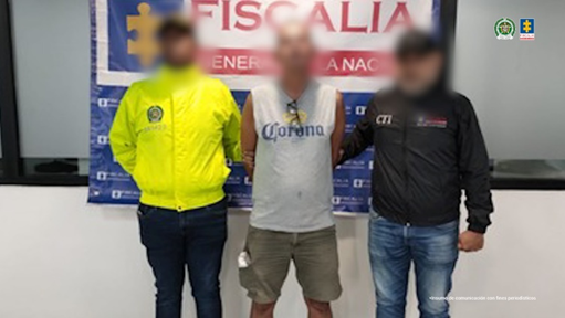 Se visualiza al capturado con el rostro difuminado, junto a un uniformado de la Policía Nacional e investigador del CTI. Detrás de ellos el banner que identifica a la Fiscalía General de la Nación.