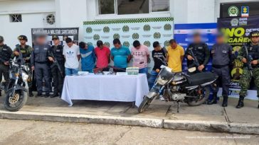 en la fotografia se visualiza los7 miembros del grupo delincuencial, entre 6 funcionarios del ejercito y el CTI de la Fiscalia, en frente de ellos se encuentra una mesa con evidencia que se les incauto al igual que una motocicleta, atras de ellos se ve un baking de la policia, del ejercito y la fiscalia